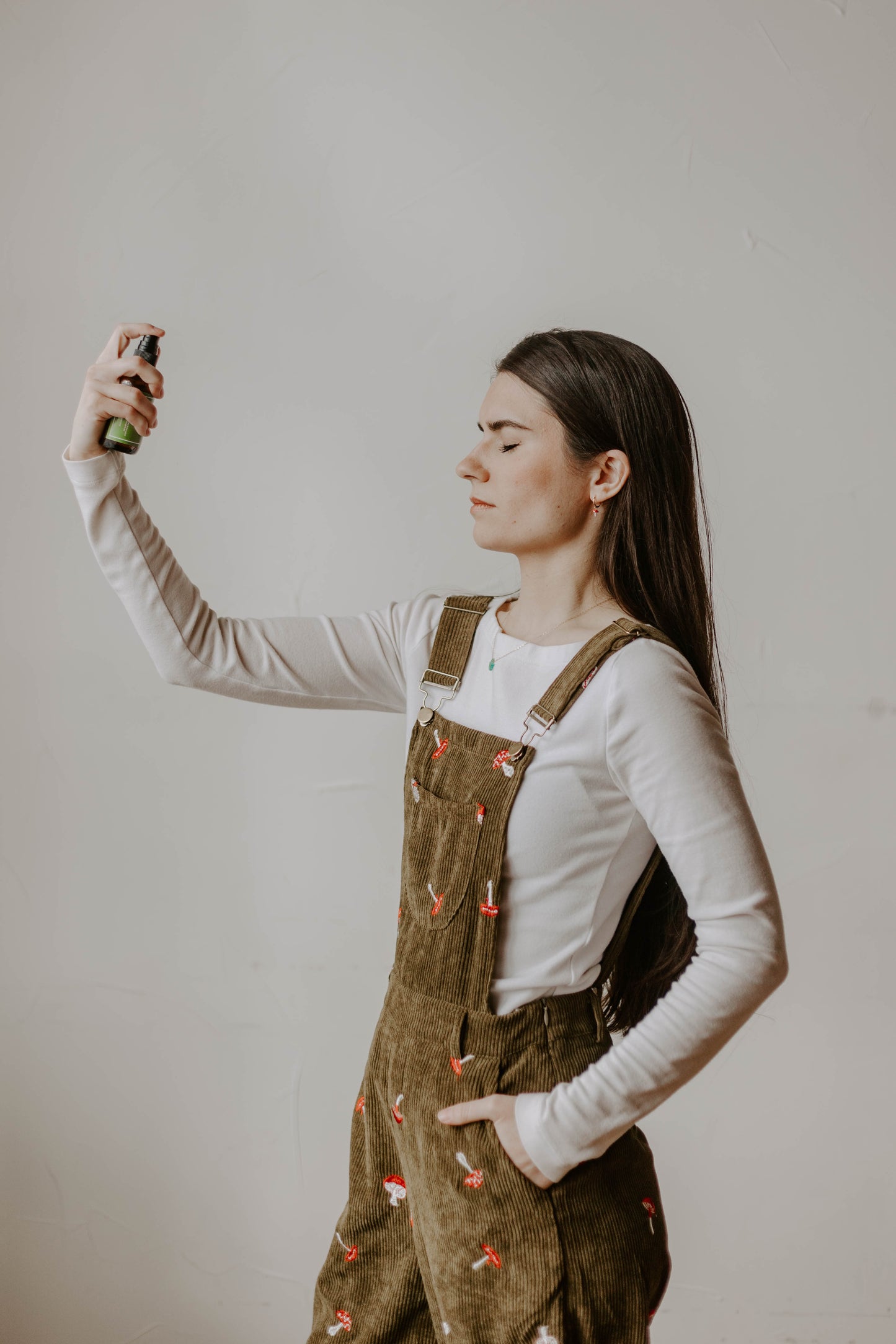 girl with long brown hair and white shirt with brown overalls that have mushrooms on them, spraying a facial toner/hydrosol on her face
