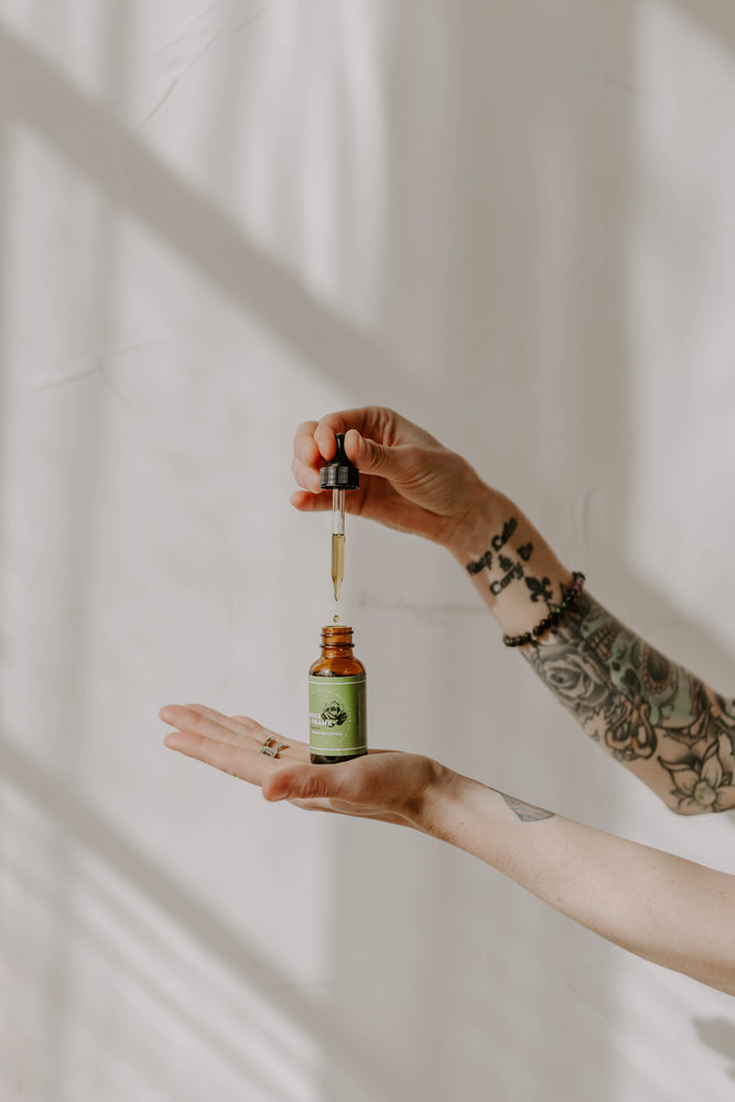 
                  
                    arm with tattoos, crystal bracelet, sunshine, facial oil, glow oil, green label, rose + frank
                  
                